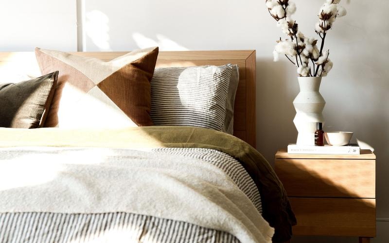 swath of sunlight shines across a well made bed and bedside table with a vase of cotton plants